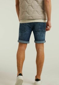 Shorts de mezclilla en azul oscuro, con puños enrollados, que cuentan con bolsillos traseros con un diseño cosido. Llevados con un chaleco acolchado beige.