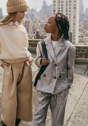 Twee kinderen staan op een dak. Eén draagt een crème sweatshirt met rhinestones, terwijl de ander een glanzend zilveren dubbel-breasted pak aan heeft.