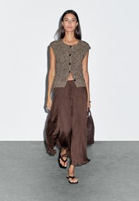 Cardigan cropped en maille marron avec des boutons, associé à une longue jupe plissée marron. Le modèle tient un sac en osier marron et porte des sandales marron.