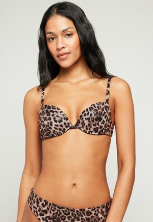 Junge Frau mit langen dunklen Haaren, die einen Leopardenmuster-BH und passende Unterteile trägt, steht vor einem schlichten hellen Hintergrund.