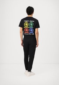 Svart t-shirt med färggrafisk design som visar kronor och lejon på baksidan, kombinerad med svarta joggers och vita skor.