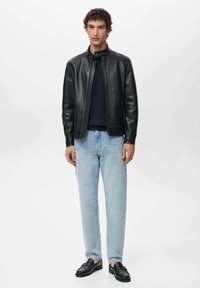 Giacca di pelle nera con colletto alto, maglione blu navy sotto, jeans azzurri e mocassini neri. Uomo in piedi contro uno sfondo semplice.