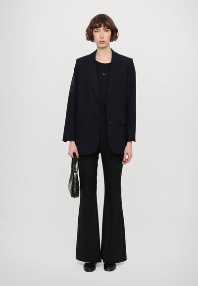 Blazer noir à double boutonnage avec une coupe ajustée, associé à un pantalon noir évasé et un sac à main vert texturé. Tissu lisse, lignes épurées.