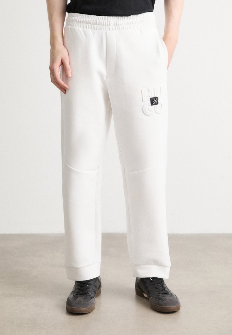 Weiße Jogginghosen mit einem elastischen Bund, seitlichen Taschen und einem strukturierten Logodetail. Die Bündchen sind eng anliegend und das Material wirkt weich.