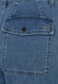 Jeans i denim med blåtvätt, med en bakficka som har en svart knapp och en broderad etikett "IVY" i guldtråd. Synlig sömdetalj.