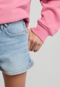 Light blue denim shorts with a frayed hem, paired with a pink fleece sweatshirt. Hand rests in the right pocket of the shorts.