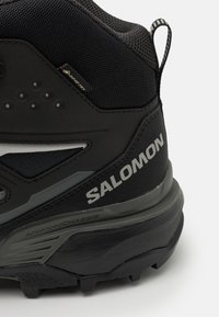 Musta vaelluskengä, jossa on verkkopinta tekstuuripinnalla, vahvistettu kanta ja harmaalla "Salomon"-merkillä, suunniteltu kestävyys ja tuki mielessä.