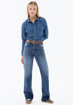 Blue denim shirt with two chest pockets, paired with high-waisted wide-leg jeans. Includes a brown belt and brown sneaker-style shoes.