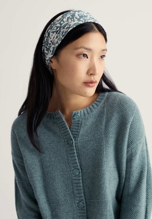 Turquoise gebreid vest met een gestructureerd patroon, voorzien van een ronde halslijn en twee blauwe knopen. Het model draagt een patroon hoofdband in turquoise en wit.