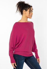 Lange ärmelige, schulterfreie magentafarbene Pullover mit lockerer Passform, gerippten Bündchen und weichem Material, kombiniert mit dunkelblauen Jeans.