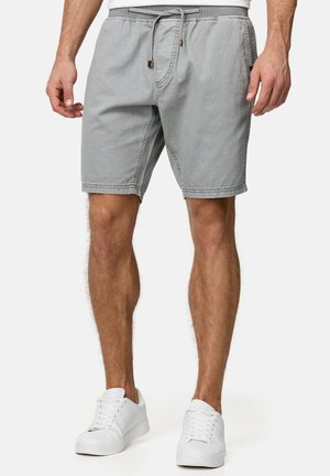 Homme portant un short gris clair avec cordon et des baskets blanches, debout devant un fond uni.