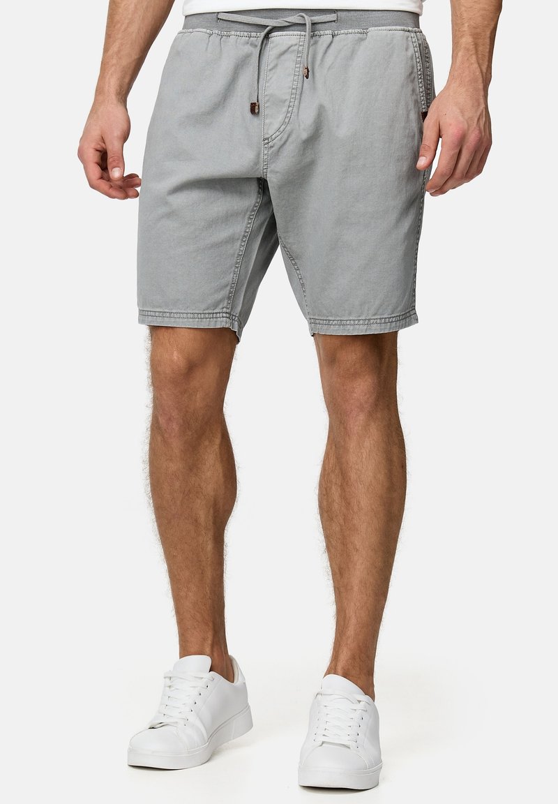 Homme portant un short gris clair avec cordon et des baskets blanches, debout devant un fond uni.