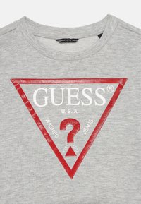 Camiseta de algodón gris con un logo triangular rojo que presenta "GUESS U.S.A." y las palabras "WASHED" y "JEANS", con un diseño de signo de interrogación central.