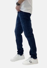 Vaqueros oscuros de ajuste slim, hechos de denim, combinados con zapatillas blancas que cuentan con detalles en negro y rojo.