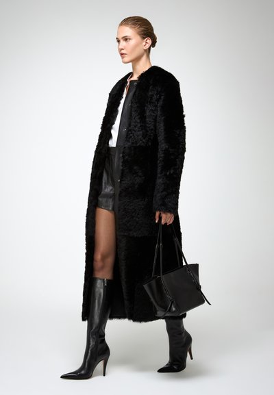 Cappotto lungo in pelliccia sintetica nera con pannello liscio nero a contrasto; abbinato a pantaloni in pelle, stivali a punta nera con tacco a stiletto, e una borsa nera.