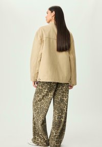 Kvinde med langt, glat hår iført en beige jakke, leopardmønstrede vide bukser, hvide sneakers og guld hoops øreringe, vendt væk.