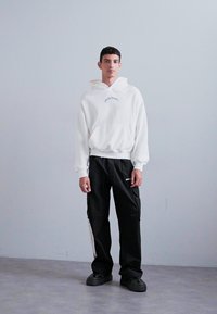 Weißer Kapuzenpullover mit einer Fronttasche und besticktem Text, kombiniert mit schwarzen Cargo-Hosen mit seitlichen Streifen und flachen schwarzen Schuhen.