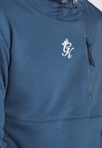 Pullover blu scuro con colletto alto, caratterizzato da un logo bianco, tasca con zip sul petto, coulisse elastiche e una texture liscia.