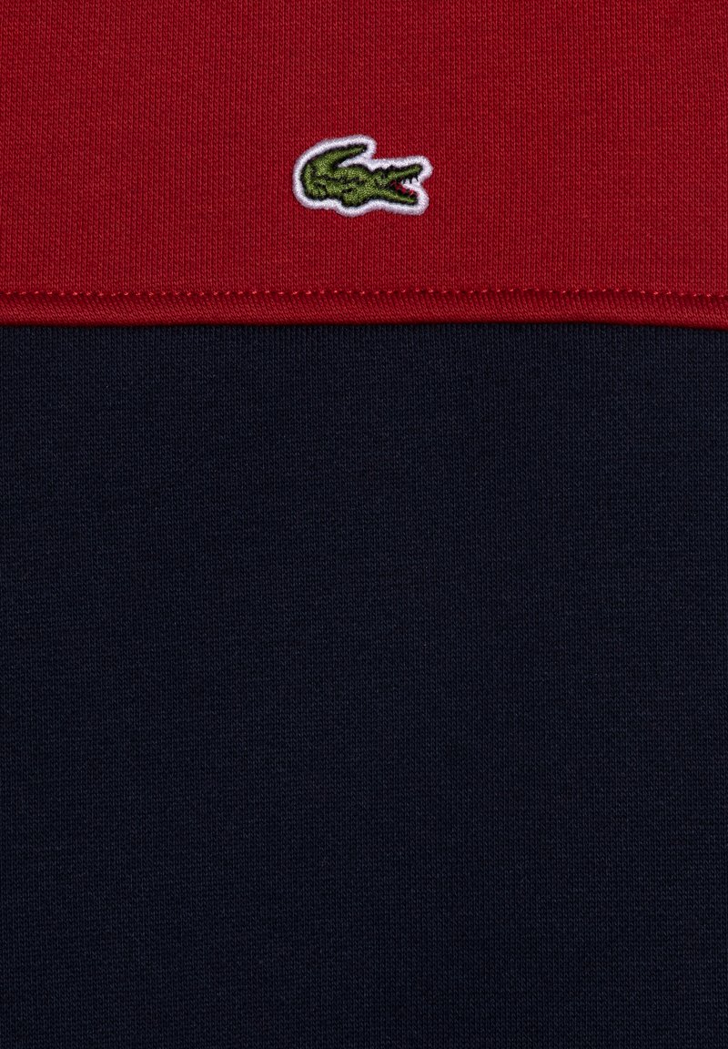 Polo shirt bicolore rouge et marine, avec un tissu en tricot texturé et un petit logo de crocodile brodé en vert sur la poitrine.