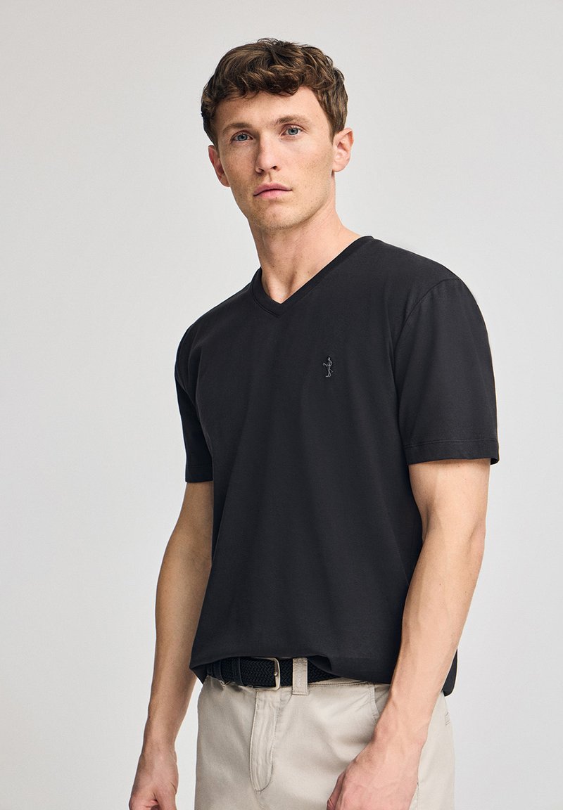 Camiseta de algodón negra con cuello en V y mangas cortas, con un logo sutil en el lado izquierdo del pecho, combinada con pantalones beige claros y un cinturón negro.