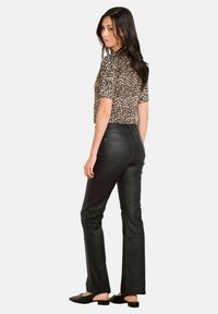 Žena stojí, kouká přes rameno, na sobě má krátký rukáv s leopardím potiskem, černé kalhoty z umělé kůže a černé slip-on boty.