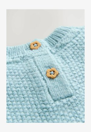 Světle modrý pletený top s texturou a knoflíkovou légu, který má jako výrazné akcenty dva dřevěné knoflíky ve tvaru květiny.