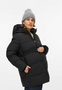 Žena u crnoj jakni s punjenjem i sivom kapom, s jednom rukom u džepu, a drugom oslonjenom na prednji dio jakne.