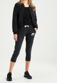 Černá bomber bunda se zlatou nášivkou, černý top na ramínka, šedé zkrácené tepláky s bílým stahováním a logem Nike, doplněné černými teniskami.