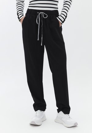 Zwarte joggingsbroek met een elastische tailleband en trekkoord, voorzien van zijzakken en een relaxte pasvorm, gecombineerd met witte sneakers.