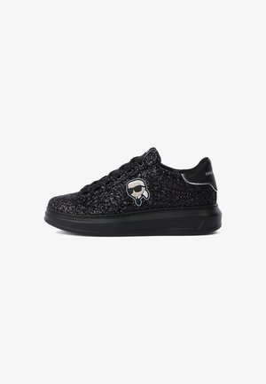 Fekete csillogós alacsonytaktikai sneaker, fűzős elülső résszel, vastag talppal és egy kis rajzos logóval, amelyen napszemüveges figura látható az oldalon.
