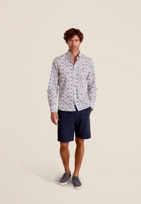 Chemise florale légère avec un fond blanc, ornée de motifs multicolores. À associer avec un short bleu marine et des chaussures grises.