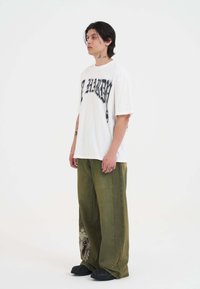 Witte oversized T-shirt met zwarte grafische tekst, gecombineerd met losse olijfgroene jeans met een ingewikkeld zijontwerp. Zwarte schoenen.