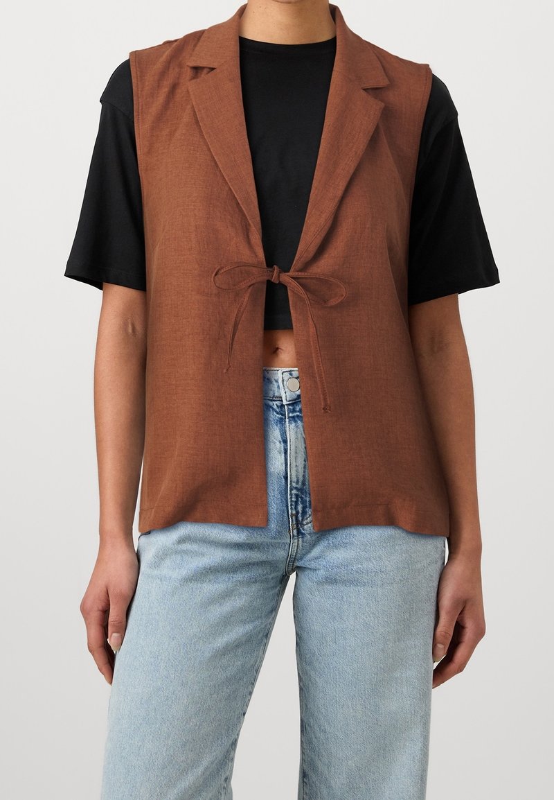 Personne portant un gilet sans manches marron noué devant, par-dessus un crop top noir à manches courtes et un jean taille haute bleu clair.