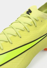 Jasnožlté futbalové topánky s textúrovaným sieťovým zvrškom, čiernymi akcentmi a lesklým neonovo zeleným prúžkom. Obsahujú šnurovanie a oranžovú podrážku.