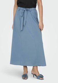 Jupe maxi bleu clair avec une taille à nouer, mettant en valeur des plis pour la forme. Associée à des sandales à talons ouvertes avec un accent floral.
