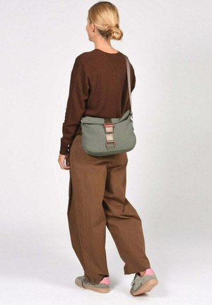 Vrouw met blond haar in een knot, draag een bruine trui en broek, draagt een grijze schoudertas met beige riem, loopt naar links.