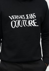 Sort langærmet sweatshirt med hvidt forvrænget tekst "VERSACE JEANS COUTURE" på brystet, båret af en person med den ene hånd i lommen.