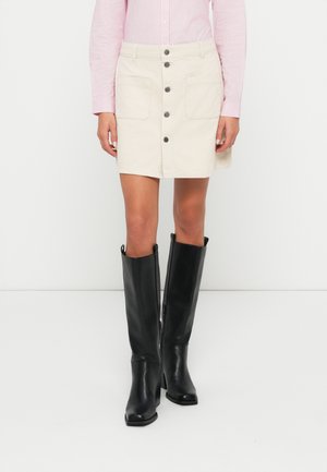 Femme portant une jupe crème boutonnée sur le devant, une chemise rose à manches longues rayée, et des bottes noires montantes, debout sur un fond blanc.