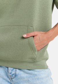 Sudadera verde oliva con bolsillo frontal, dobladillo y puños acanalados, y una textura suave. Llevada sobre un denim azul claro con un acabado desgastado.