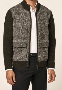 Uomo che indossa una giacca in lana texturata nera e bianca con zip sopra una camicia bianca con bottoni e pantaloni neri.