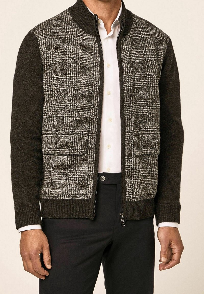Uomo che indossa una giacca in lana texturata nera e bianca con zip sopra una camicia bianca con bottoni e pantaloni neri.