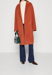 Donna che indossa un cappotto lungo color ruggine, un maglione bianco, jeans blu e stivali a punta marroni, mentre tiene una borsa verde con un flap testurizzato.