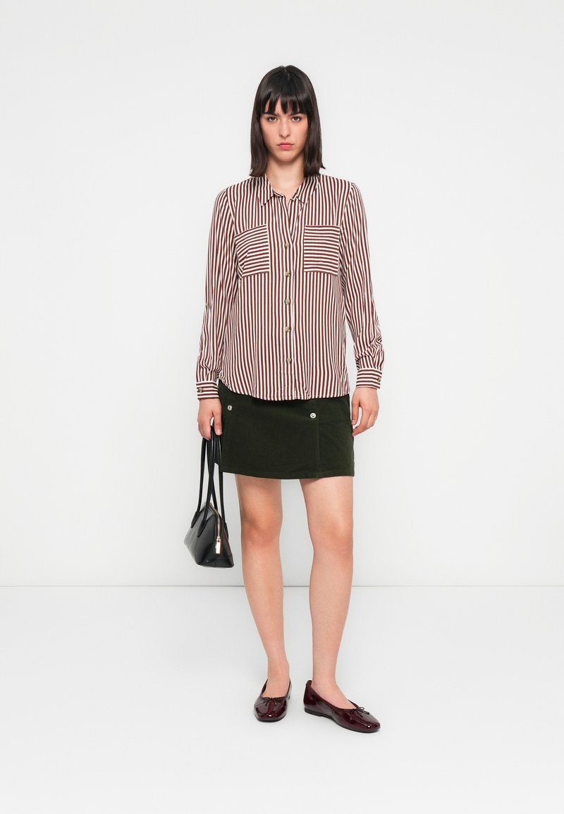 Chemise à rayures rouge et blanche, jupe en corduroy verte avec détails en boutons, sac à main noir et ballerines en vernis bordeaux.