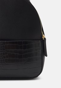 Sac noir avec une partie supérieure en cuir lisse et un motif crocodile texturé à la base, avec des accessoires en or et une fermeture à zip.