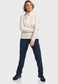 Hellbeige Fleecejacke mit hohem Kragen, Reißverschluss und seitlichen Taschen, kombiniert mit dunkelblauen Hosen und schwarzen Wanderschuhen.
