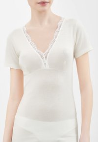 Top bianco a maniche corte con scollatura a V profonda, caratterizzato da una delicata rifinitura in pizzo lungo il colletto. Tessuto morbido e liscio con una forma aderente.