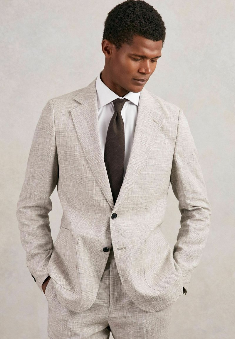 Traje de lino gris claro con una textura tejida, dos botones y bolsillos laterales. Combinado con una camisa blanca y una corbata marrón oscuro.