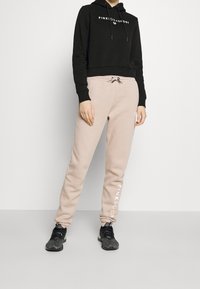 Schwarzer kultivierter Hoodie mit weißem Logo, kombiniert mit hellbeigen Jogginghosen mit seitlichem Branding. Schwarze Sportschuhe runden den Look ab.