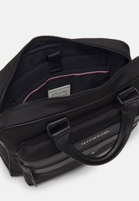 Sort nylon taske med struktureret overflade, der har en lynlås lukning, to håndtag og flere indvendige lommer beklædt med sort tekstil.