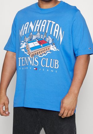 Hombre con camiseta azul con gráfico del Manhattan Tennis Club, horizonte de la ciudad y logo de Tommy Jeans.